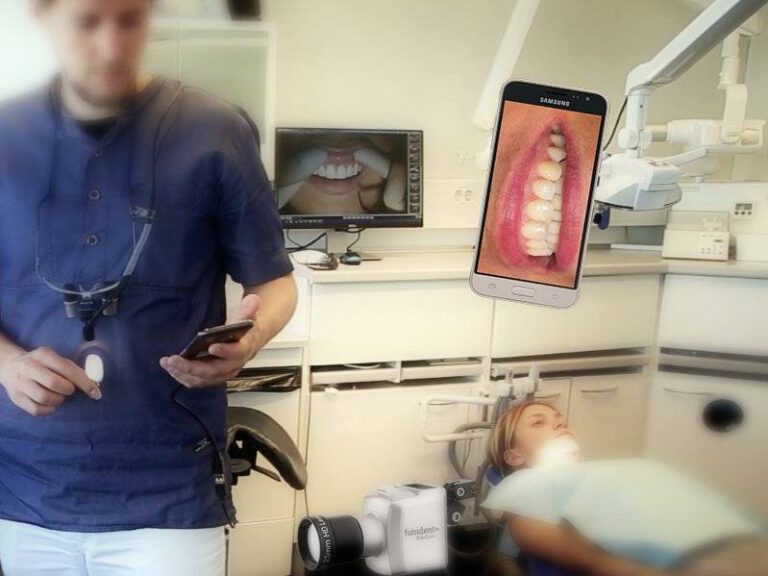 Ein Mann in blauer medizinischer Kleidung steht in einer Zahnarztpraxis und bedient ein Smartphone. Im Hintergrund ist ein Bildschirm mit einem Bild eines Mundes zu sehen. Im Vordergrund sieht man eine Patientin auf einem Zahnarztstuhl und eine Aufnahme von Zähnen auf einem weiteren Smartphone-Bildschirm.
