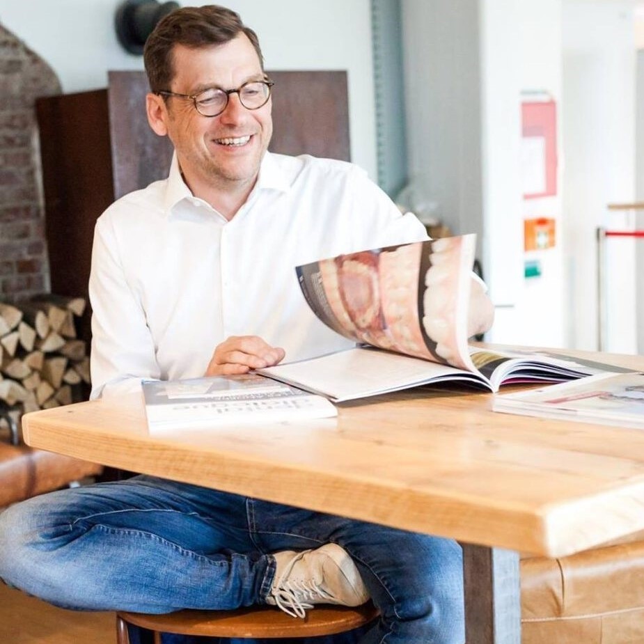 Mann mit Brille und weißem Hemd sitzt lächelnd an einem Tisch und blättert in einem Magazin. Auf dem Tisch liegen weitere aufgeschlagene Bücher mit Bildern von Zähnen.
