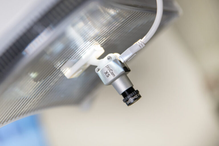 Eine kleine, silberne Kamera mit der Aufschrift "proCam XS", die an einer medizinischen Lampe montiert ist. Ein weißes Kabel ist an die Kamera angeschlossen.